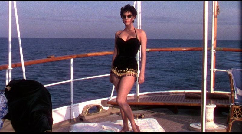 Ava Gardner stands on the deck of a yacht wearing sunglasses and a bathing suit.